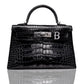 A black handbag with a crocodile pattern and a silver initial 'B' attached to it, set on a reflective surface, collection-preview.