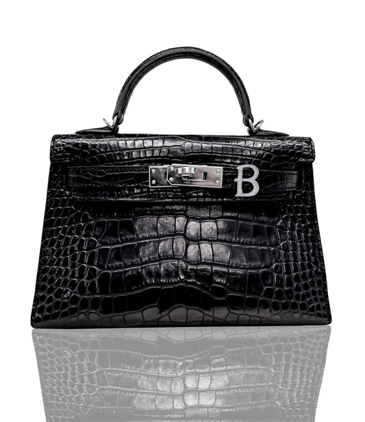 A black handbag with a crocodile pattern and a silver initial 'B' attached to it, set on a reflective surface, collection-preview.
