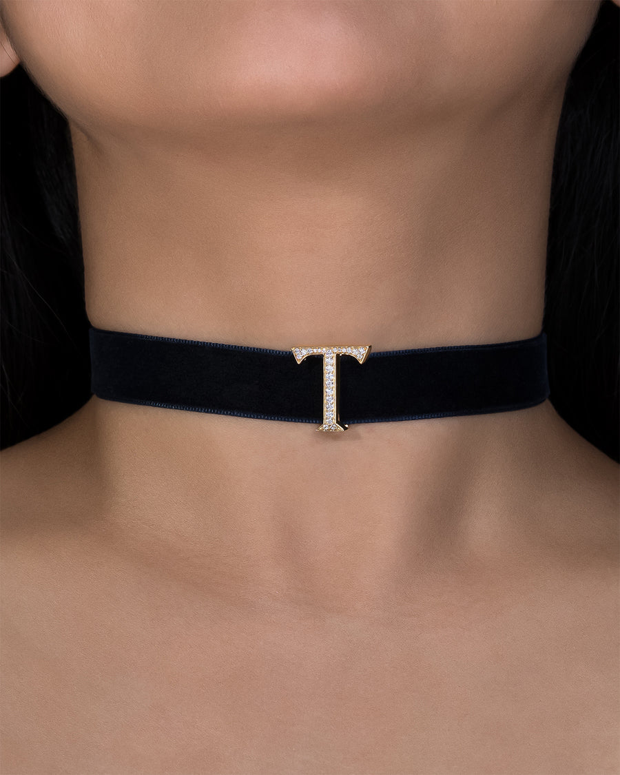 Black velvet choker with a gold 'T' pendant on a person wearing a black top.